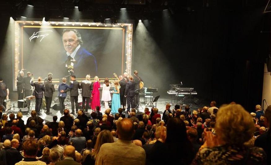 Kuvassa on etualalla yleisöä selin seisomassa ja katsoo näyttämölle. Lavalla on mies- ja naisesiintyjiä seisomassa selin ja katsoo suurta Kari Tapion muotokuvaa, jossa Tapiolla on sininen takki päällään.