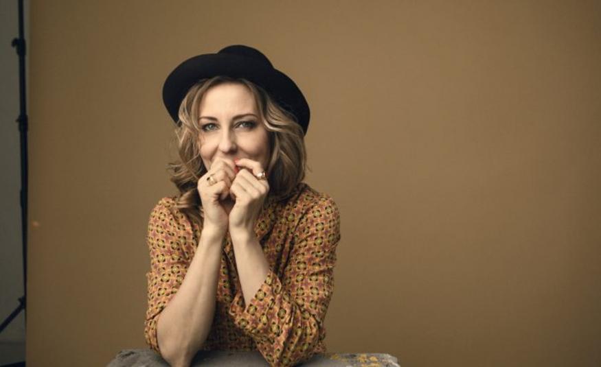 Kuvassa on Mariska puolivartalokuvassa ja hänellä on musta hattu päässään sekä kellertävä, pikkukuvioinen pusero päällään. Hänellä on molemmat kädet kevyesti nyrkissä suun edessä. Tausta on ruskehtavaa seinää.