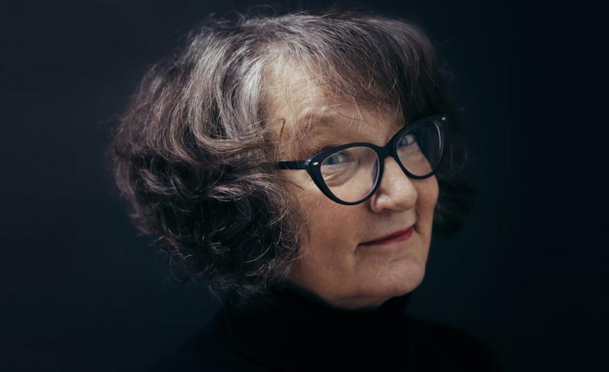 Kuvassa on Monika Fagerholmin kasvokuva ja hänellä on kihartavat tumman harmaat hiukset  sekä mustasankaiset silmälasit. Tausta on musta.