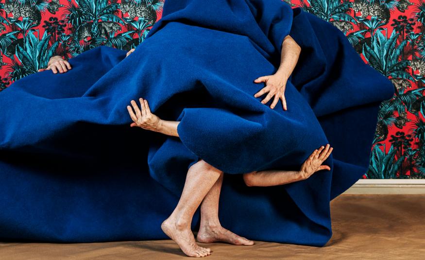 Kuvassa on sinisen kankaan alla henkilöitä ja käsiä näkyy kankaan välistä ja keskellä alhaalla on kävelevät jalat. Taustalla on kukallista puna-turkoosi-mustaa tapettia.