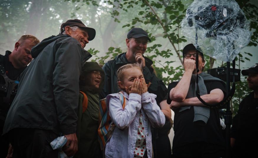 Kuvassa on Siiri Virtanen keskellä vaalean sinisessä takissa. Ympärillä on aikuisia miehiä ja naisia tummissa takissa ulkona lehtipuiden alla. 