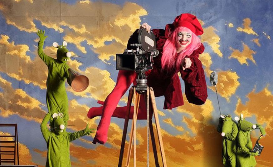 Kuvassa taustalla kelta-harmaita pilviä sinisellä taivaalla. Vasemmalla on kaksi vihreätä hiireksi pukeutunutta hahmo ja toinen seisoo okapäillä. Keskellä on vanhanaikainen filmikamera ja siinä päällä roikkuu nainen punaisissa saappaissa legginsseissä sekä viininpunaisessa takissa. Takana on pieninä kaksi vihreätä hiirtä.