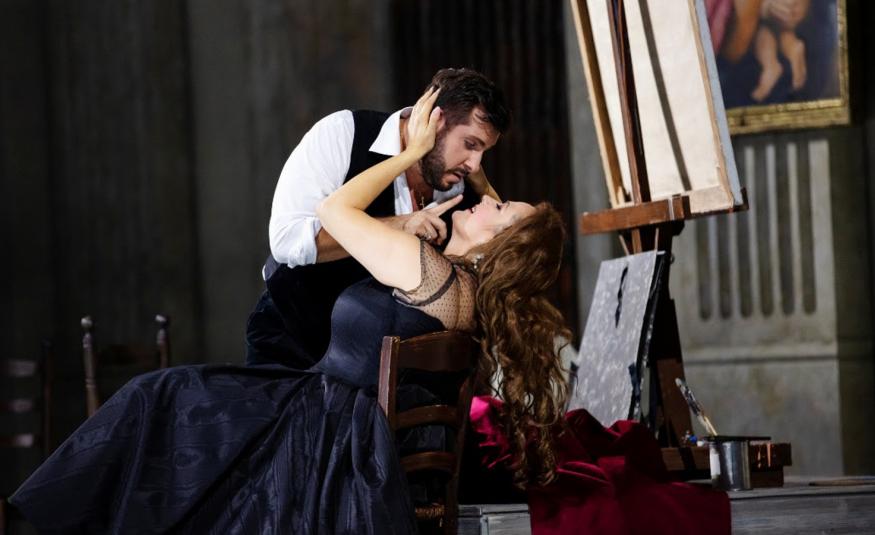 Kuvassa on Toscan roolin laulaja tumman sinisessä pitkässä puvussa istumassa tuolissa ja nojaa selkänojaan.  Hänen ylläpuolellaan on kuvartuneena kasvoja kohti mies valkoisessa puserossa ja mustassa liivissä. Tausta on tumma.