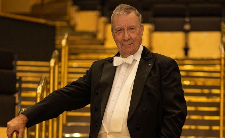 Kuvassa on Okko Kamu seisomassa portaikossa, jossa on metalliset kaiteet. Hänellä on frakki päällään ja pitää oikeata kättään kaiteella. Kuvan sävy on ruskehtava.