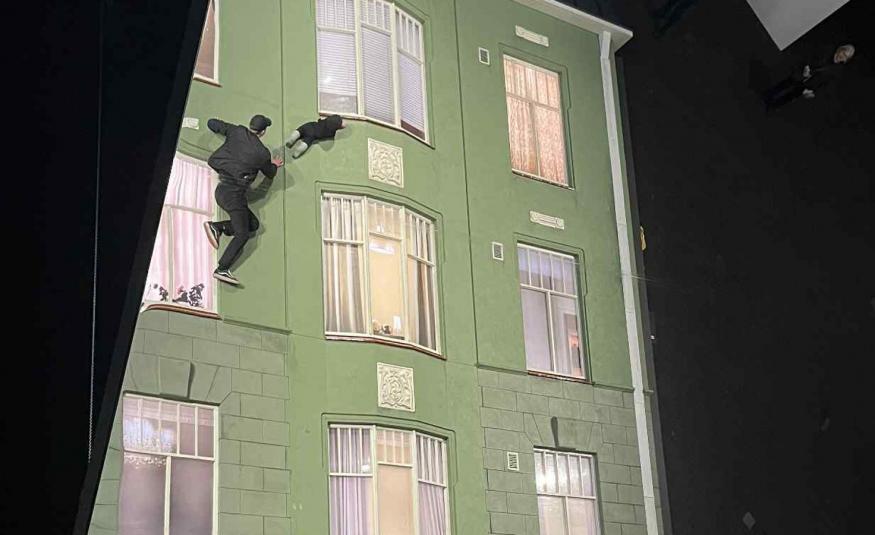 Kuvassa on molemmin puolin mustaa ja keskellä on vaalean vihreä vanha kerrostalo, jossa on kolmeen osaan jakautuneet ikkunoita kolmessa rivissä. Vasemmalla ylhäällä on nuori mies mustassa puvussa mukanaan vauva kiipeämässä seinällä.