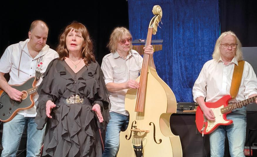 Kuvassa on vasemmalta kitaristi ja hänen vieressään edessään nainen ruskeassa rimpsutetussa mekossa ja keskellä on vaaleaa bassoa soittava mies. Oikealla on kitaraa soittava mies valkoisessa paidassa ja farkuissa.