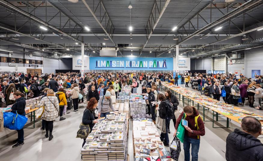 Kuvassa on isossa hallissa useita pitkiä pöytiä täynnä kirjoja ja niitä katselemassa on paljon ihmisiä. Takana on iso sininen lakana, jossa on valkoisin kirjaimin Ilmaiskirjapäivä.