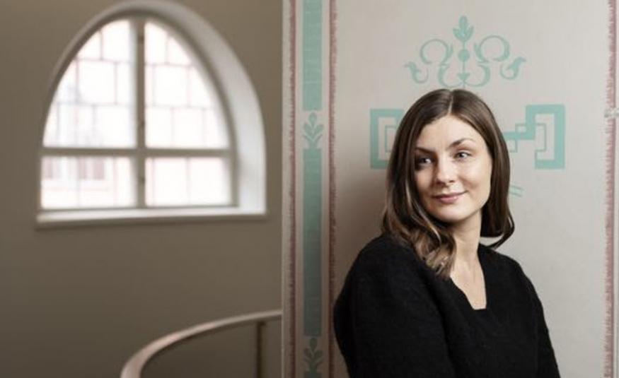 Kuvassa on oikealla Iida Turpeinen puolivartalokuvassa mustassa puserossa. Hän on tilassa, jossa on vaalea, turkoosikoristeinen seinämän reunus. Taaempana näkyy kaarevaa ikkunaa.