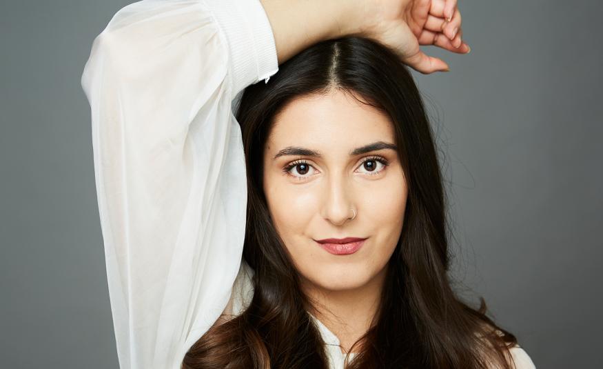 Kuvassa on Iris Candeleria puolivartalokuvassa ja hänellä on pitkt tummat hiukset. Hänellä on valkoinen pitkähihainen pusero ja hän pitää oikeaa kättään päälaellaan. Tausta on harmaa.