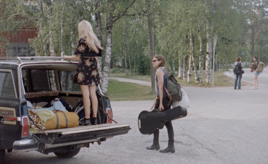 Kuvassa on vasemmalla auton takakontti auki ja sen päällä seisoo reunalla selin Alina Tomnikov. Taustalla on koivumetsää ja asfaltilla oikealla kävelee poispäin nuori mies.