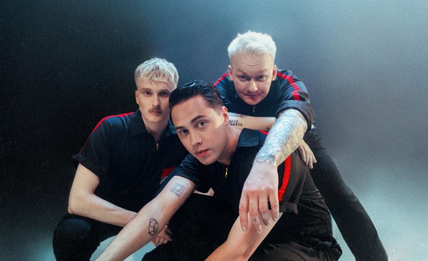 Kuvassa ovat Kuumaa-yhtyeen kolme jäsentä istumassa tiiviisti yhdessä. Heillä on tummat t-paidat päällään. Tausta on tumman sininen.