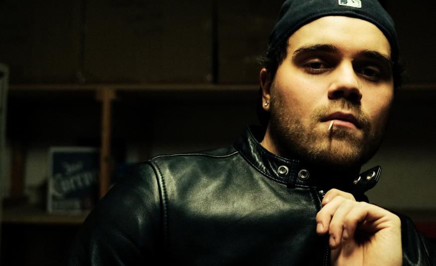 Kuvassa on oikeassa reunassa puolivartalokuvassa ja hänestä näkyy mustalla pohjalla kasvot ja vasenta kämmentä. Hänellä on musta nahkatakki päällään.