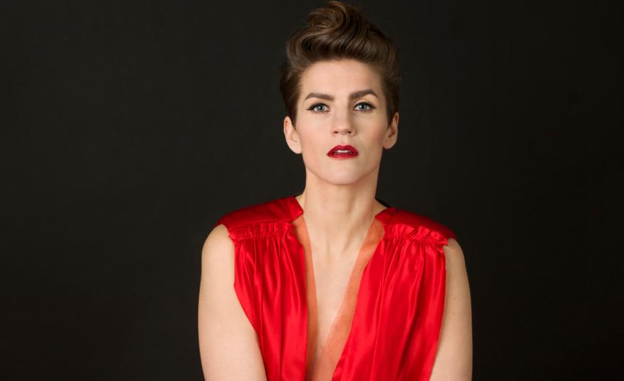 Kuvassa on Maria Ylipää istumassa puolivartalokuvassa. Hänellä on punainen, hihaton mekko päällään ja siinä on v-kaula-aukko. Tausta on tumma.