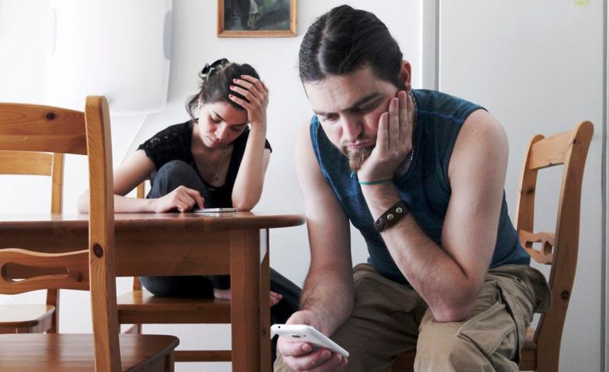 Kuvassa on etualalla oikealla nuori mies istumassa, nojaa vasemmalla kädellään poskeensa ja katsoo oikeassa kädessään olevaa kännykkää. Pöydän takana istuu nuori nainen ja katsoo myös kännykkäänsä. 