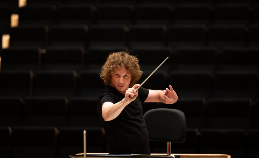 Kuvassa on Santtu-Matias Rouvali johtamassa orkesteria tahtipuikko silmiensä tasalla puolivartalokuvassa. Hänellä on musta t-paita päällään. Tausta on musta.