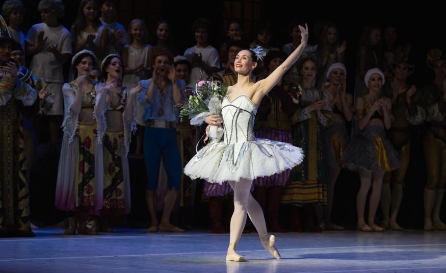 Kuvassa on keskellä valkoisessa balettipuvussa Sofia Gustafsson ja hänellä on vasen käsi ojennettuna sekä kukat oikeassa kädessään. Taustalla näkyy himmeästi muita esiintyjiä.