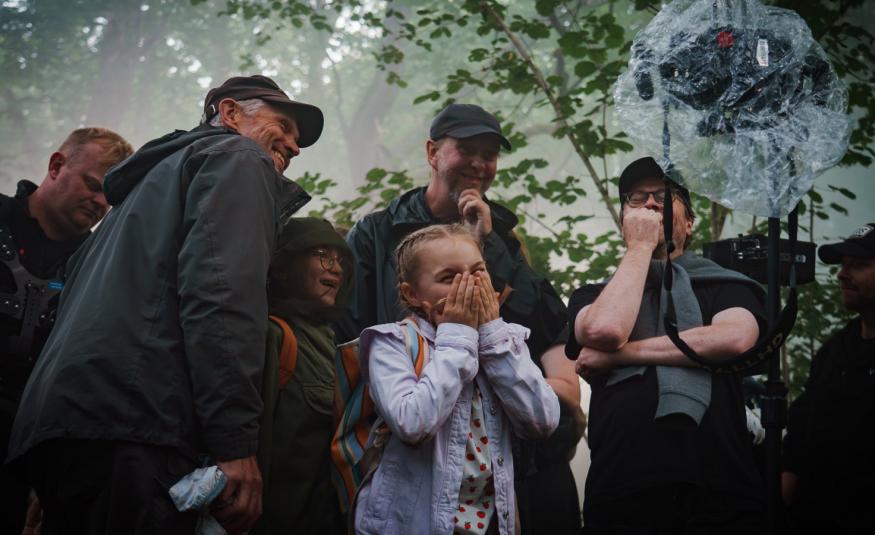 Kuvassa on keskellä vaalean sinisessä farkkutakissa Siiri Virtanen ja hänen ympärillään on kuvausryhmän kuusi henkilöä tummissa ulkovaatteissa. Yläpuolella on vihreitä lehtipuun oksia ja kaikki seisovat puun alla.