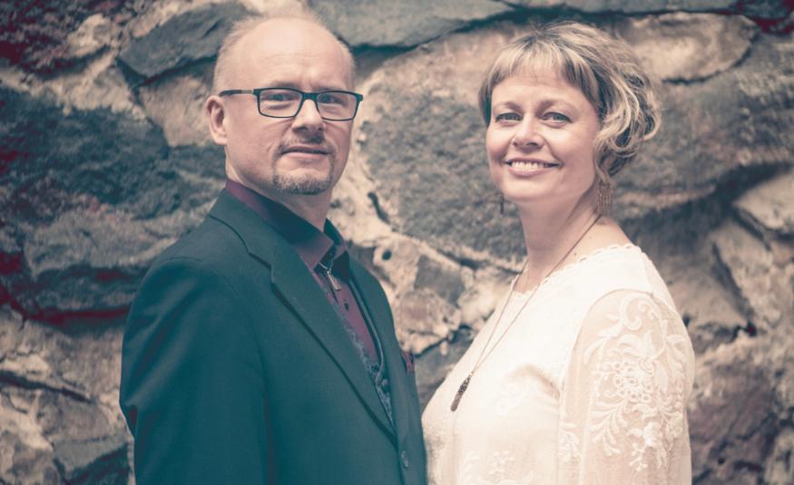 Kuvassa on vasemmalla Tapio Lappalainen  mustassa takissa ja Sanna Lappalainen vaaleassa puserossa. Taustalla on kiviseinää, jossa on suuria, tummia kiviä ja vaaleita välimuurauksia. 