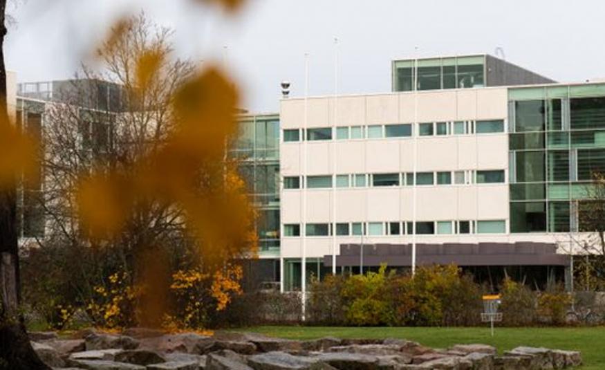  Kuvassa on vasemmalla olevan lähes lehdettomän puun takana oleva rakennus, joka on L.n muotoinen ja jonka oikea osa on vaalea. Alla näkyy litteähköjä kiviä ja pihapensaita