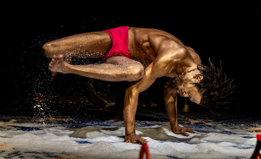 Kuvassa mies, jolla on pronssiseksi maalattu iho, ja hän käsien päällä ja muu keho vinottain vaaka-asennossa ylhäällä. Hänellä on punaiset alushousut. Tausta on musta ja lattialla on valkoista vaahtoa.