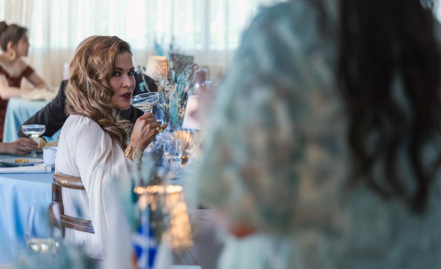 Kuvassa on ravintolatila ja oikealla edessä on himmeästi seisomassa nainen, josta näkyy turkoosin valkoista puseroa ja hieman pitkiä hiuksia selässä. Vasemmalla kauempana istuu Jenni Vartiainen sivuttain ja hän katsoo tulijaa sekä siemaileen oikeassa kädessään olevaa drinkkiä.