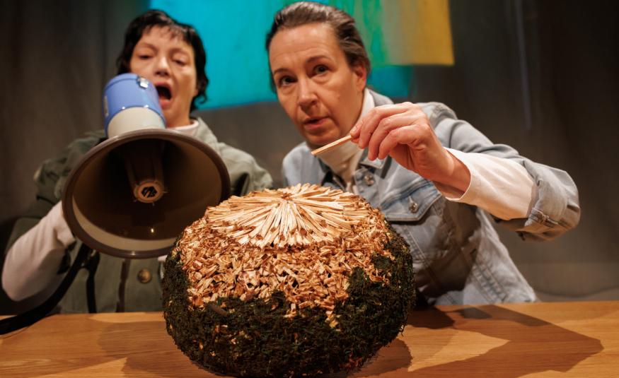 Kuvassa ovat istumassa pöydän ääressä Teija Töyry kädessään megafoni ja Heidi Syrjäkari ojentamassa vasenta kättään kohti pöydällä olevaa isoa palloa. Se on ruskea ja siinä on kultausta päällä. 