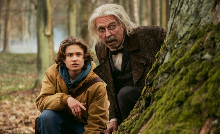 Kuvassa ovat vasemmalla metsässä kyykyssä suuren puunrungon vieressä Kuura Rossi ja hänen vieressään vähän ylempänä Martti suosalo pitkissä vaaleissa hiuksissa. Taustalla näkyy puunrunkoja.