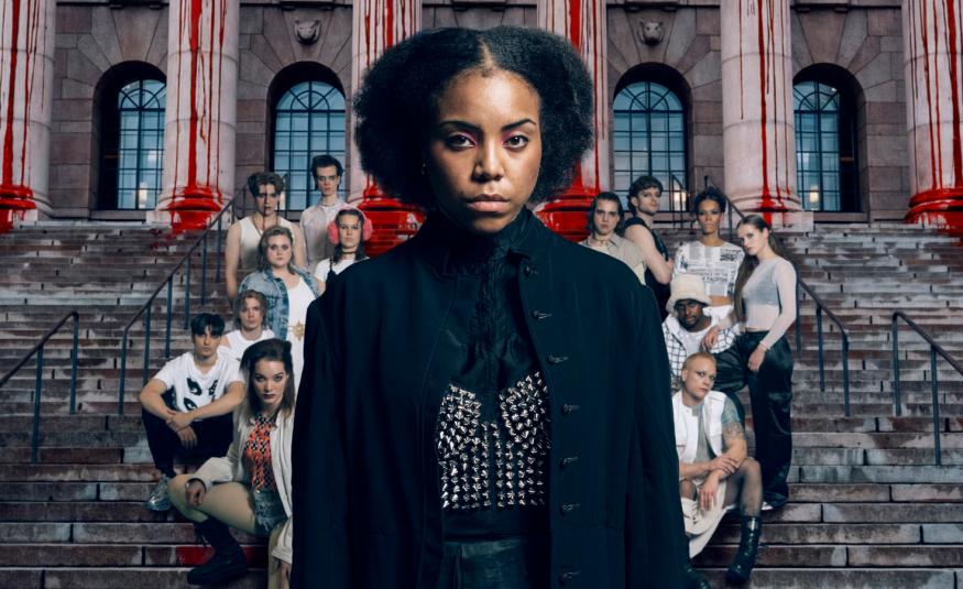 Kuvassa on keskellä mustassa mekossa Abdirahman Bile iso kokoisena ja hänen takanaan on joukko muita näyttelijäitä ryhmänä rapuilla punertavan tiilirakennuksen edessä. 