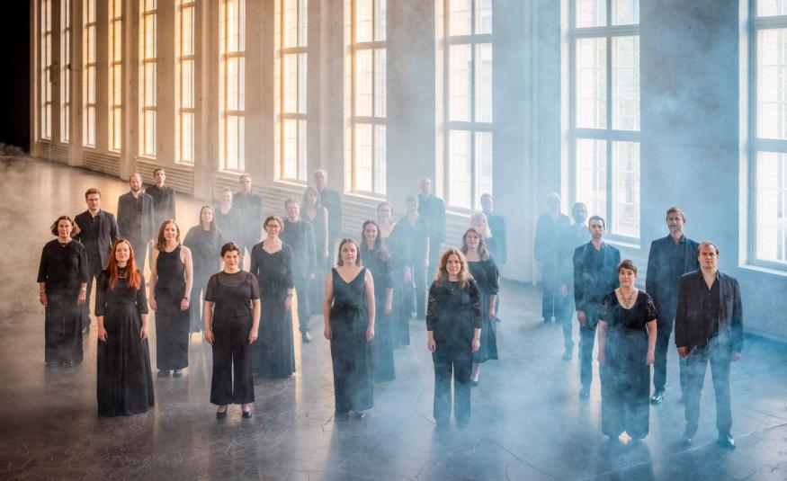 Kuvassa on Helsingin kamarikuoron jäsenet seisomassa mustissa mekoissa ja puvuissa. He ovat isossa hallissa, jossa on korkeat ruutuikkunat rivissä, joista tulee valoa.  