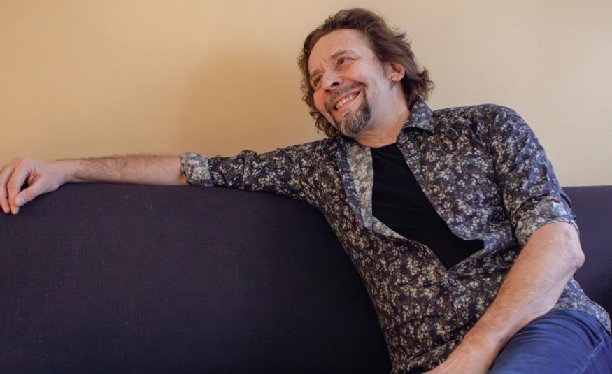 Kuvassa on Ilari Johansson istumassa mustalla sohvalla ja hänen oikea kätensä on ojentuneena sohvan selkänojan päällä. Hänellä on mustan kirjava paita päällään. Seinä on kellertävä.