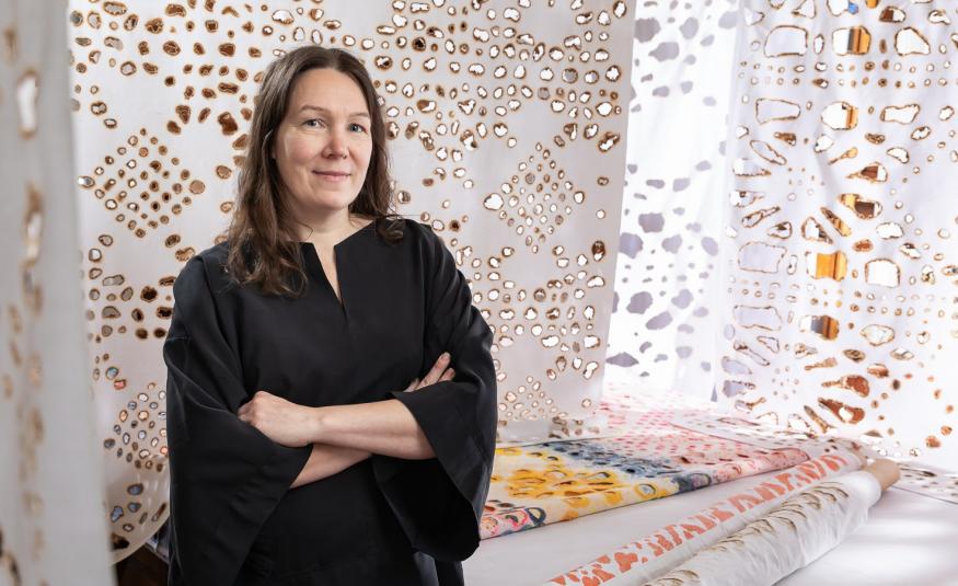 Kuvassa on vasemmalla Johanna Pöykkö mustassa mekossa puolivartalokuvassa. Taustalla on ripustettu kankaita, joissa on ruskeareunaisia reikiä erilaisina kuvioina. 