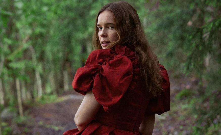 Kuvassa on Marketta Tikkanen selin kävelemässä metsätietä, jonka varrella on koivuja. Tkkasella on punainen mekko päällään ja siinä on puhvihihat.