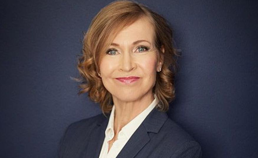 Kuvassa on Leena Niemistö ja hän on puolivartalokuvassa. Hänellä on tumman sininen jakku ja valkoinen paitapusero päällään sekä puolipitkät, kiharat punertavat hiukseton. Tausta on tumman sininen. 