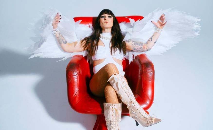 Kuvassa on Mira Luoti istumassa punaisessa kiiltävässä nojatuolissa. Hänellä on valkoinen mekko ja kädet levitettyinä sivuille ja niissä on enkelin siivet. Tausta on vaalea ja siinä on varjo vasemmalla.