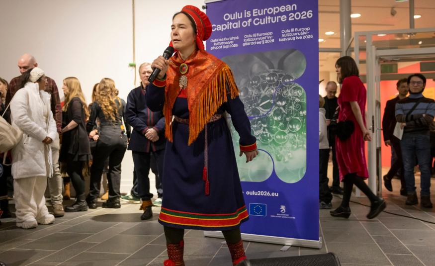 Kuvassa on Anna Morottaja keskellä kuvaa esiintymässä Lapin puvussa, joka on tumman sininen ja siihen kuuluu puna-oranssi -kuvioinen huivi ja päähine.  Taustalla näkyy Oulu2026 roll-up ja vasemmalla yleisöä.