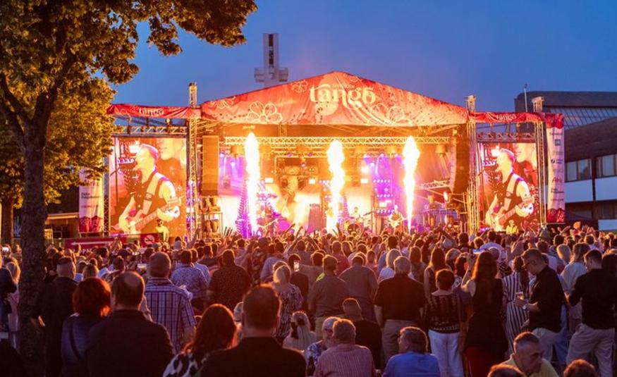 Kuvassa on takana on oranssin punainen teltta, jossa on keskellä pyrotehosteita ja vasemmalla sekä oikealla näkyy kitaristi soittamassa.  Etualalla näkyy yleisöä selin. Taustalla on kesäyön sinertävää taivasta.