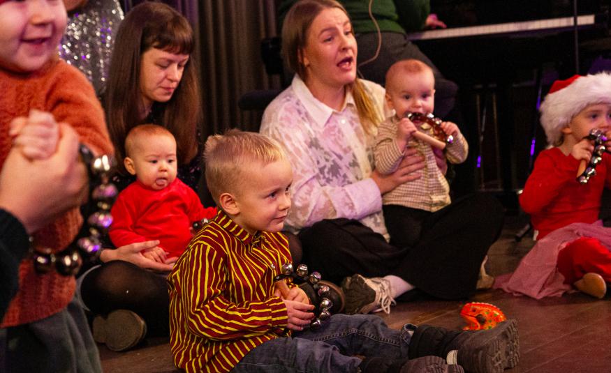 Kuvassa on äitijä ja vauvoja sekä pieniä lapsia istumassa lattialla.  He katsovat esitystä.