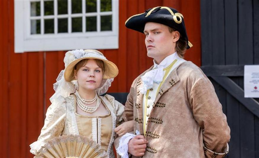 Kuvassa on vasemmalla nuori nainen vaalean ruskeassa vanhanaikaisessa mekossa ja lierihatussa. Asussa ja hatussa on valkoisia röyhelöitä ja brodyyrejä. Oikealla on nuori mies, jolla on musta aatelisen suikkahattu päässään sekä koristeellinen vaalean ruskea takki päällään ja valkoinen röyhelöpaita. Taustalla on punaisen puurakennuksen seinää ja valkoinen, pieniruutuinen ikkuna.