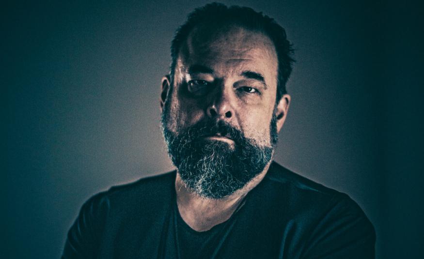 Kuvassa on A.W. Yrjänä puolivartalokuvassa ja hänellä on musta t-paita päällään ja musta parta. Kuvan tausta on tumma.