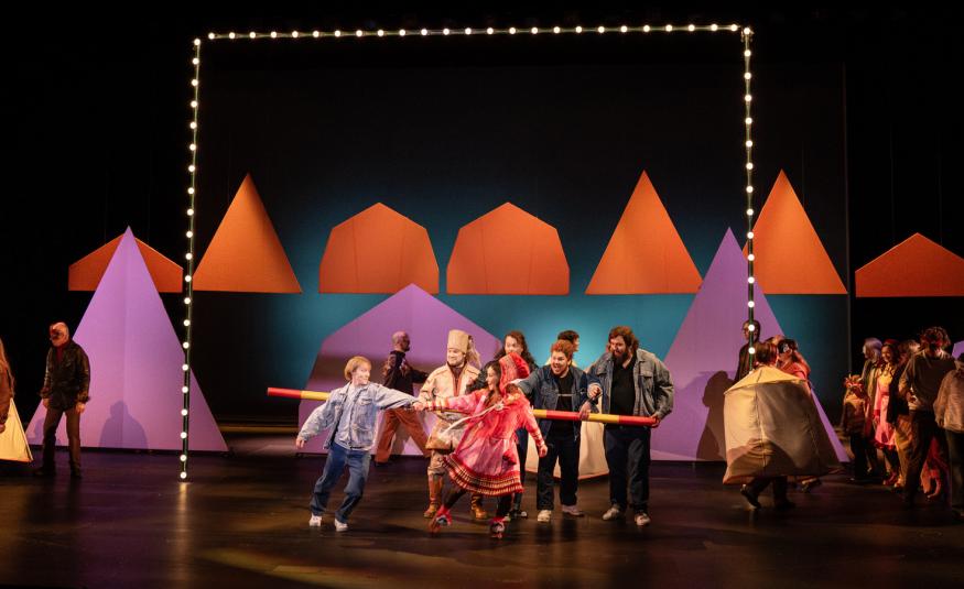 Kuvassa on tummalla näyttämöllä takana punaisia kartiomaisia telttoja. alemana on näytteljöitä tanssimassa edessä värikkäissä vaatteissa ja taaempana seisoo harmassa puvuissa olevia näyttelijöitä. Vasemmalla on kartiomainen lila teltta.