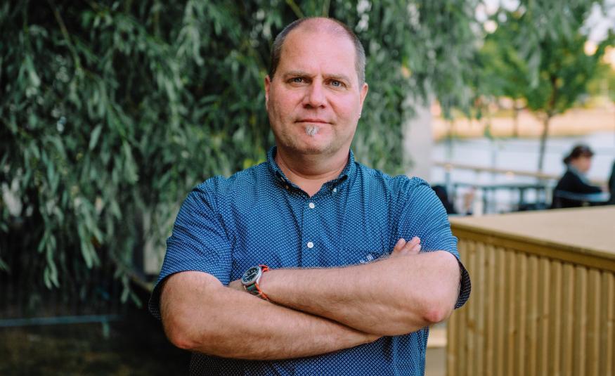 Kuvassa on keskellä Stuba Nikula seisomassa puolivartalokuvassa.  Hänellä on sininen, lyhythihainen  paita päällään ja hänellä on kädet toistensa päällä.  Taustalla on alaspäin laskeutuvia lehtipuun oksia.