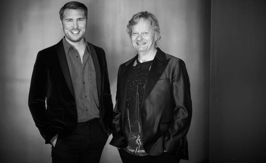 Kuvassa on seisomassa vasemmalla aarne Pelkonen mustassa takissa ja harmaassa paidassa kädet taskussa. Oikealla on Iiro Rantala mustassa kiiltävässä takissa. Tausta on tumma ja siinä on taustavaloa. Kuva on musta-valkoinen.