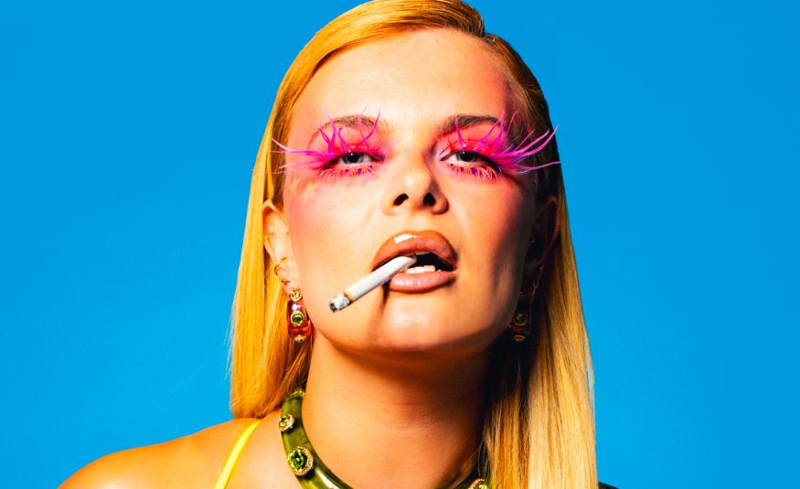 Kuvassa on puolivartalokuvassa Erika Vikman ja hänellä on pitkät vaaleat hiukset, punaiseksi meikatut silmänympärykset ja tupakka hampaissaan. Tausta on turkoosi. 