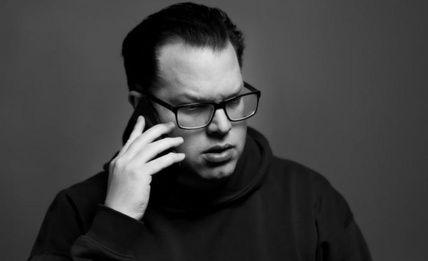 Kuvassa on Jonas Olson puolivartalokuvassa. Hänellä on oikeassa kädessään kännykkä korvallaan. Hänellä on tummasankaiset silmälasit ja musta poolokauluspusero päällään. Tausta on harmaa.