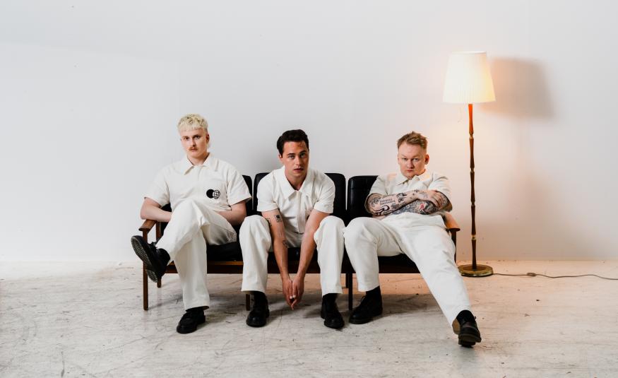 Kuvassa ovat Kuumaa-yhtyeen kolme jäsentä istumassa sihvalla. Heillä kaikilla on valkoiset vaatteet. Oikealla on jalkalamppu. Kuvan tausta on valkoinen ja lattia vaalea.