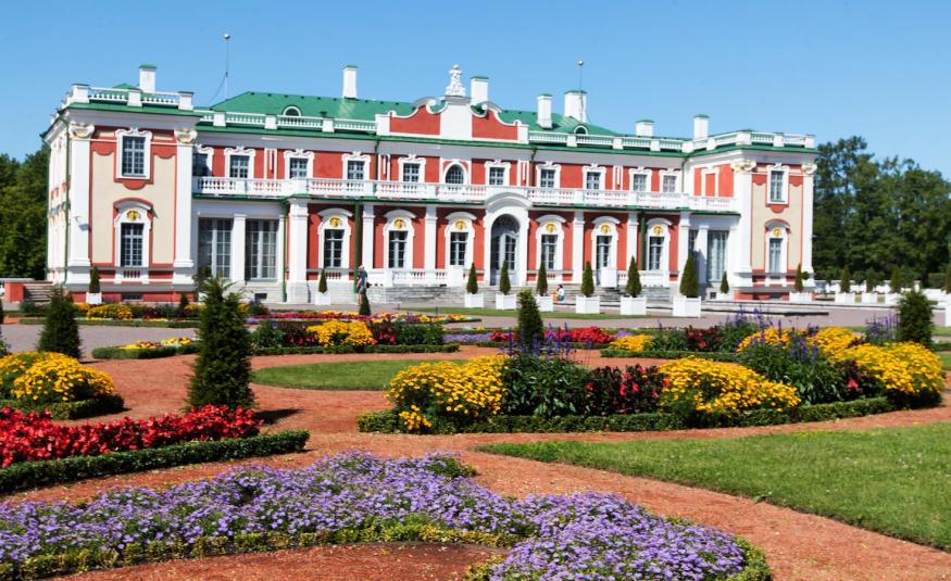 Kuvassa on Kadriorgin taidemuseon rakennus, joka on puna-valkoinen ja siinä on korkeat koristeelliset ikkunat ja valkoinen koko julkisivun yläparveke. Edessä on värikkäitä kukkaistutuksia.