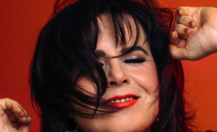 Kuvassa on Kaija Koo kasvokuvassa. Hänellä on tummat hiukset osittain kasvoilla.  Tausta ruskean punainen.