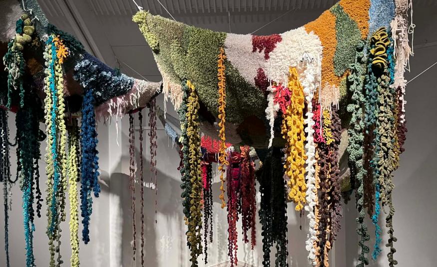  Kuvassa on naruilla roikkumassa erivärisiä kiharaisia, paksuja lankoja. Oikealla toisella narulla on yläosassa eri värisiä ryijymäisiä osia yläosassa, joista roikkuvat, paksut langat lähtevät.