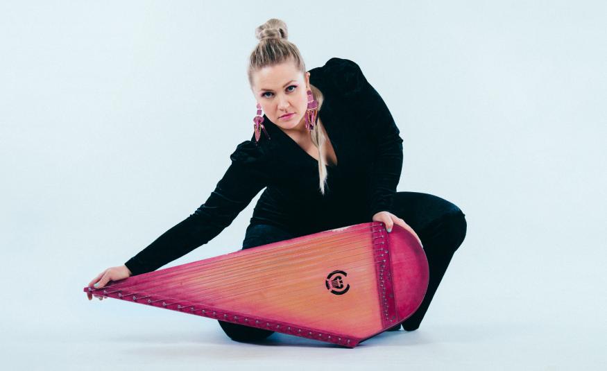 Kuvassa on Maija Kauhanen mustassa asussa kumartuneena punaisen vasemmalle suipoksi muotoillun kanteleen takana ja hän pitää oikealla kädellään kanteleen päästä kiinni. Tausta on vaalean sininen.  