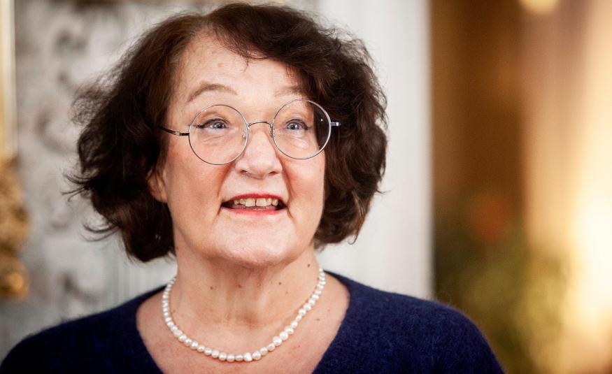 Kuvassa on Monika Fagerholm puolivartalokuvassa ja hänellä on tummat hiukset sekä pyöreät silmälasit. Hänellä on tumma pusero päällään ja valkoiset helmet.